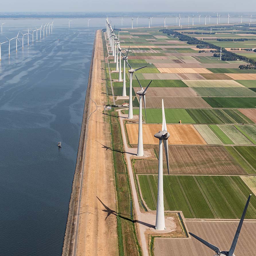 Foto com visão aérea de paisagem agrícola contornada por turbinas eólicas gigantes ao longo da costa.